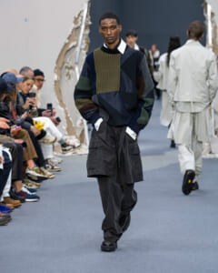 Male model walks the runway in a patchwork knit sweater and oversized black cargo pants, audience watching from the sides.