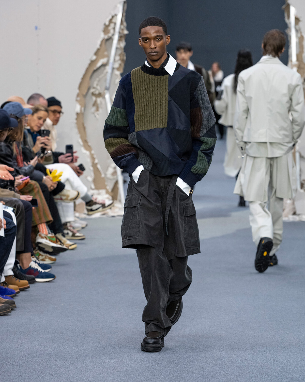 Male model walks the runway in a patchwork knit sweater and oversized black cargo pants, audience watching from the sides.