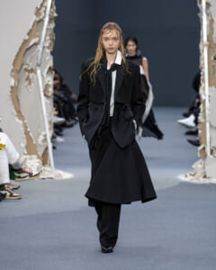 Model walks the runway in a tailored black suit with a white shirt and long coat, hands in pockets, wet hair.