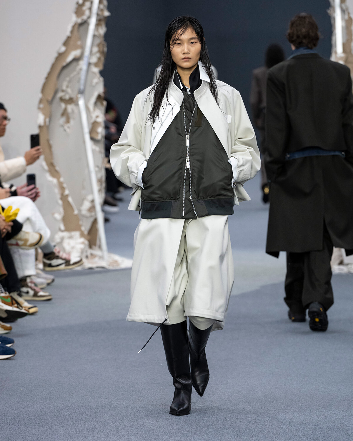 Runway model walks the catwalk in a pale oversized coat over a dark green bomber and light gray shorts, paired with black knee-high boots.