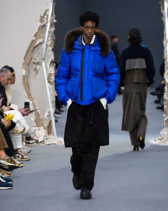 Runway model in a bright blue puffer jacket with fur-trimmed hood, paired with black layered bottoms on a fashion catwalk.