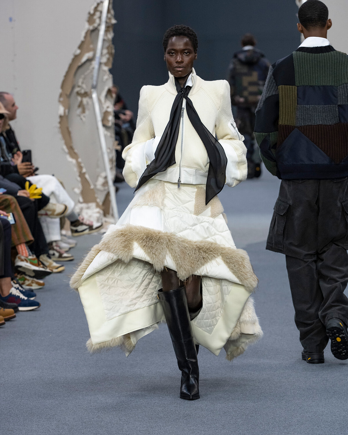 Model walks the runway in a cream quilted coat with fur trim and knee-high black boots