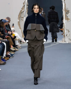 Runway model walks the catwalk in a navy ribbed turtleneck with an oversized brown waist-layered top over matching brown wide-leg trousers and black ankle boots; audience seated along the sides.