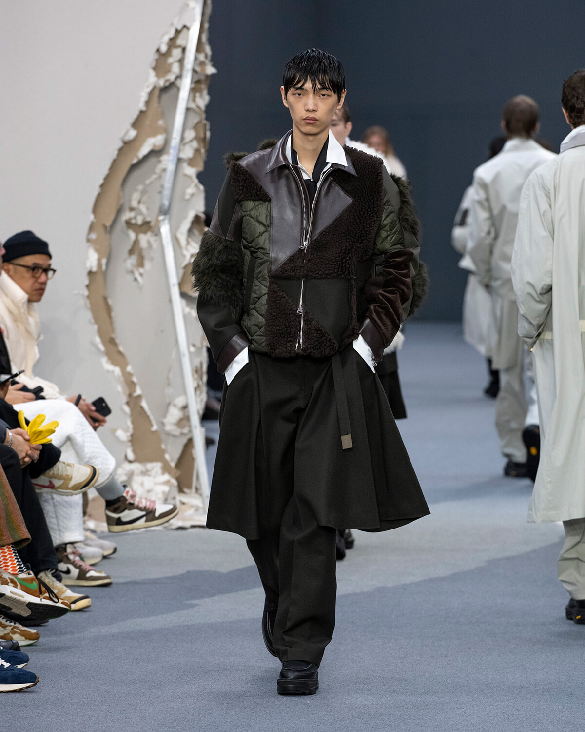 Male model walks the runway in a dark patchwork shearling jacket and wide black trousers during a fashion show.
