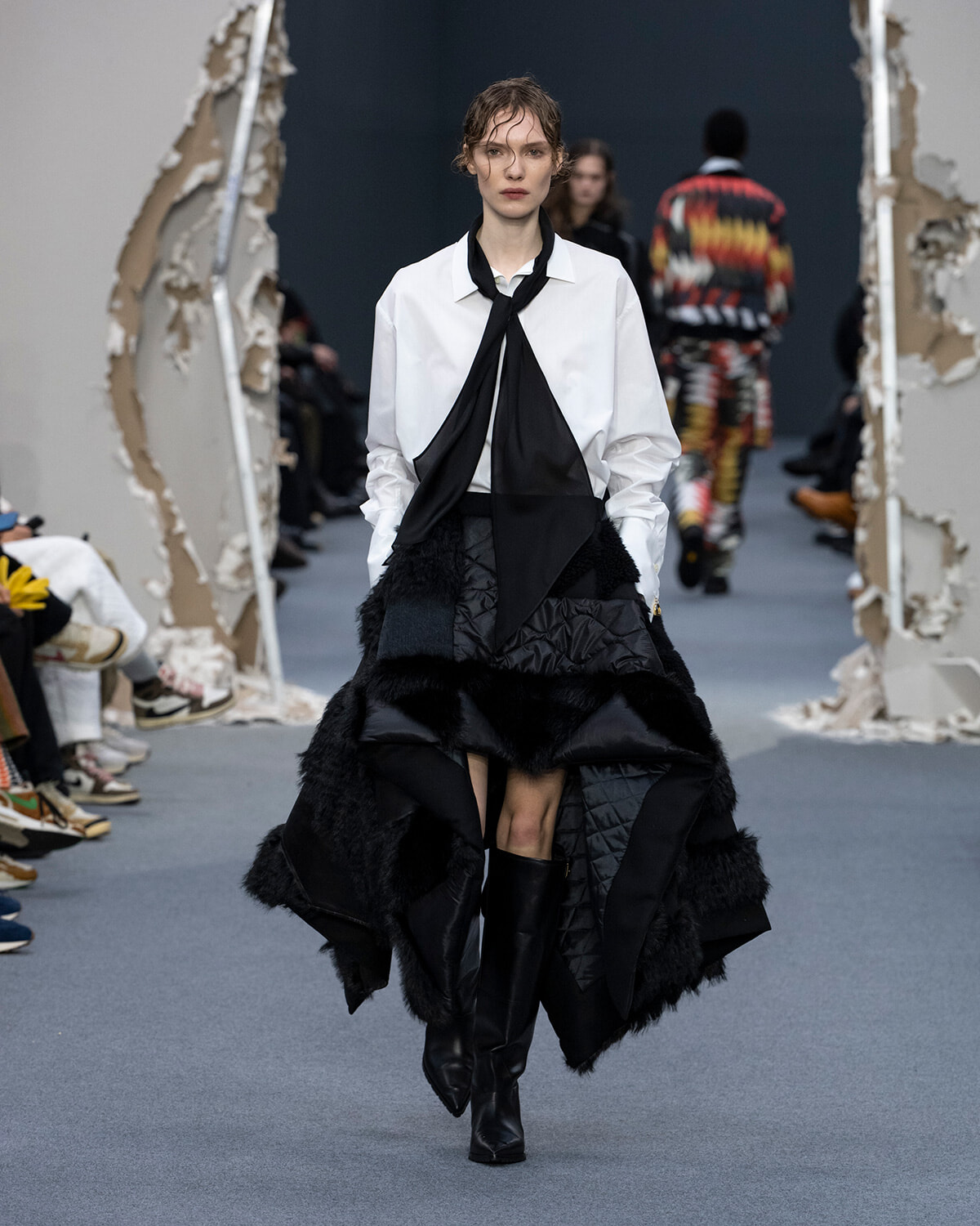 Model on a fashion runway wearing a white button-down shirt with a black scarf-tie and a voluminous black quilted skirt with fur trim, paired with knee-high black boots.
