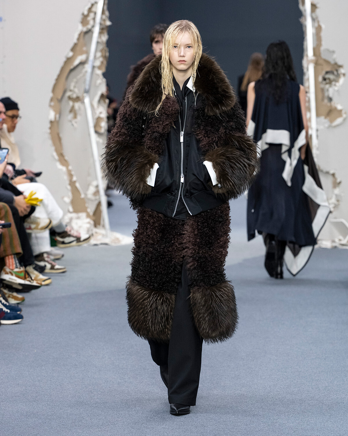 Model on a runway wearing a long dark fur coat with oversized collar and black pants, posing for the show.