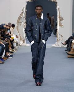 Male model walks a runway in a dark denim jacket and wide-leg jeans, burgundy shoes, audience on both sides.