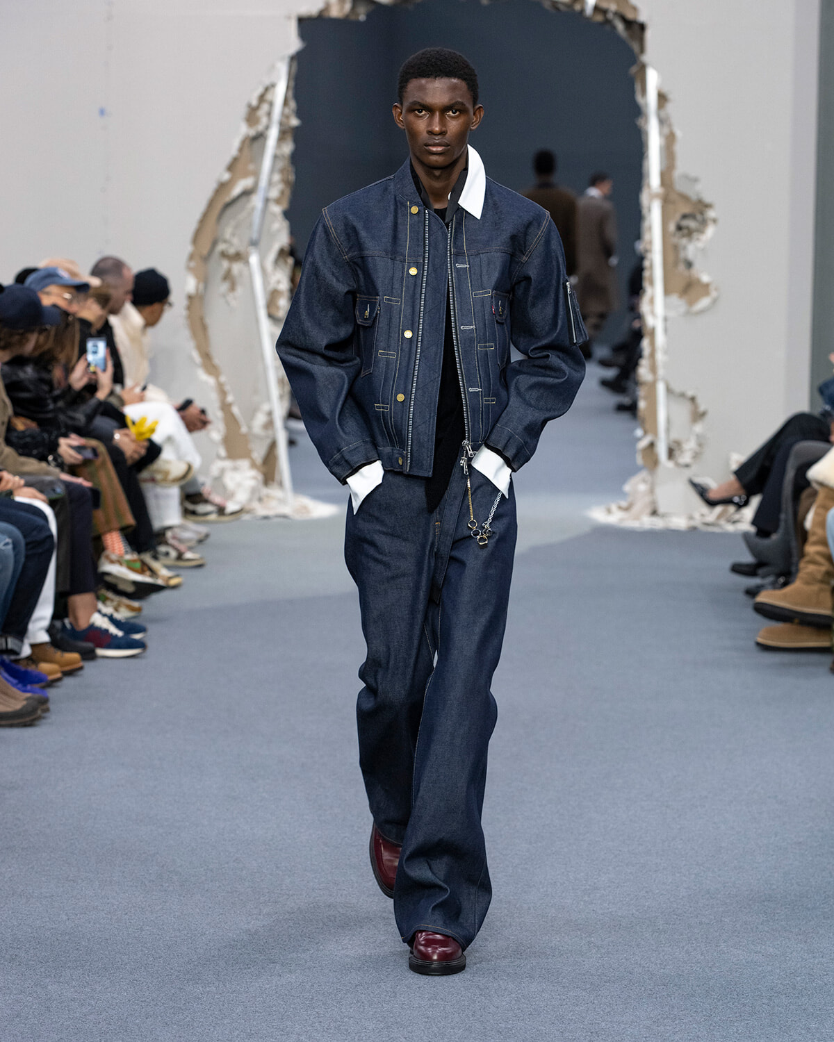 Male model walks a runway in a dark denim jacket and wide-leg jeans, burgundy shoes, audience on both sides.