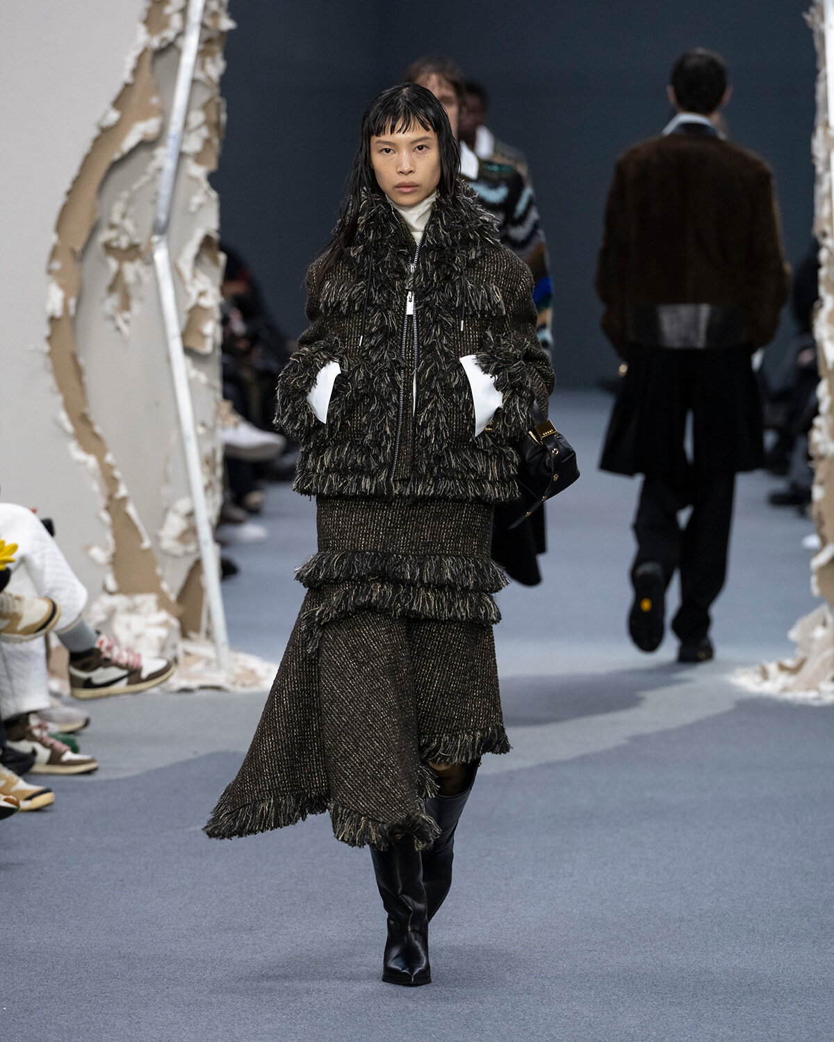 Model walks runway in a dark brown textured fur coat with white inner sleeves and a matching frayed knit skirt, hands in pockets, black boots.