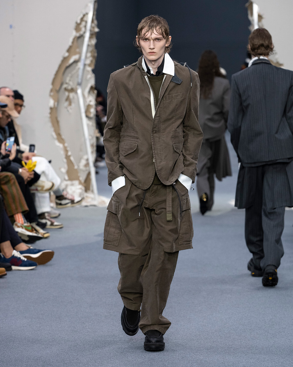Male model walks the runway in olive-brown utility jacket and cargo pants with white-collar shirt, audience on the left and textured sculpture in background.