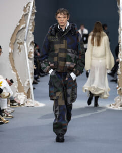 Male model walks the runway wearing a bulky quilted patchwork jacket and matching pants in muted blues and greens.