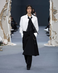 Model on a runway wearing a white button-down shirt with a black necktie scarf and black wide-leg pants/skirt combo, hands in pockets.