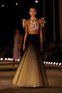 Model on a dimly lit runway wearing a sheer brown top with white floral appliqués and a high-waisted black-to-cream striped pleated skirt.