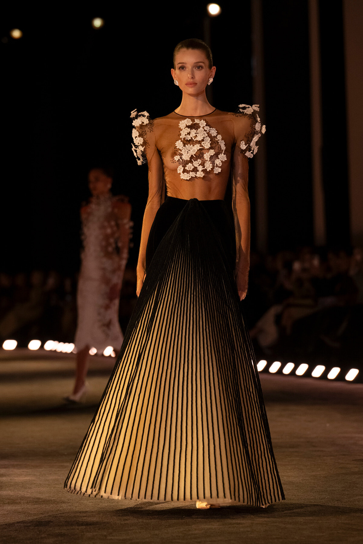 Model on a dimly lit runway wearing a sheer brown top with white floral appliqués and a high-waisted black-to-cream striped pleated skirt.
