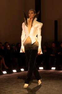 Model walks the runway in a cream tailored blazer with wing-like shoulder pieces, gradient pants, and metallic heels, audience in background.