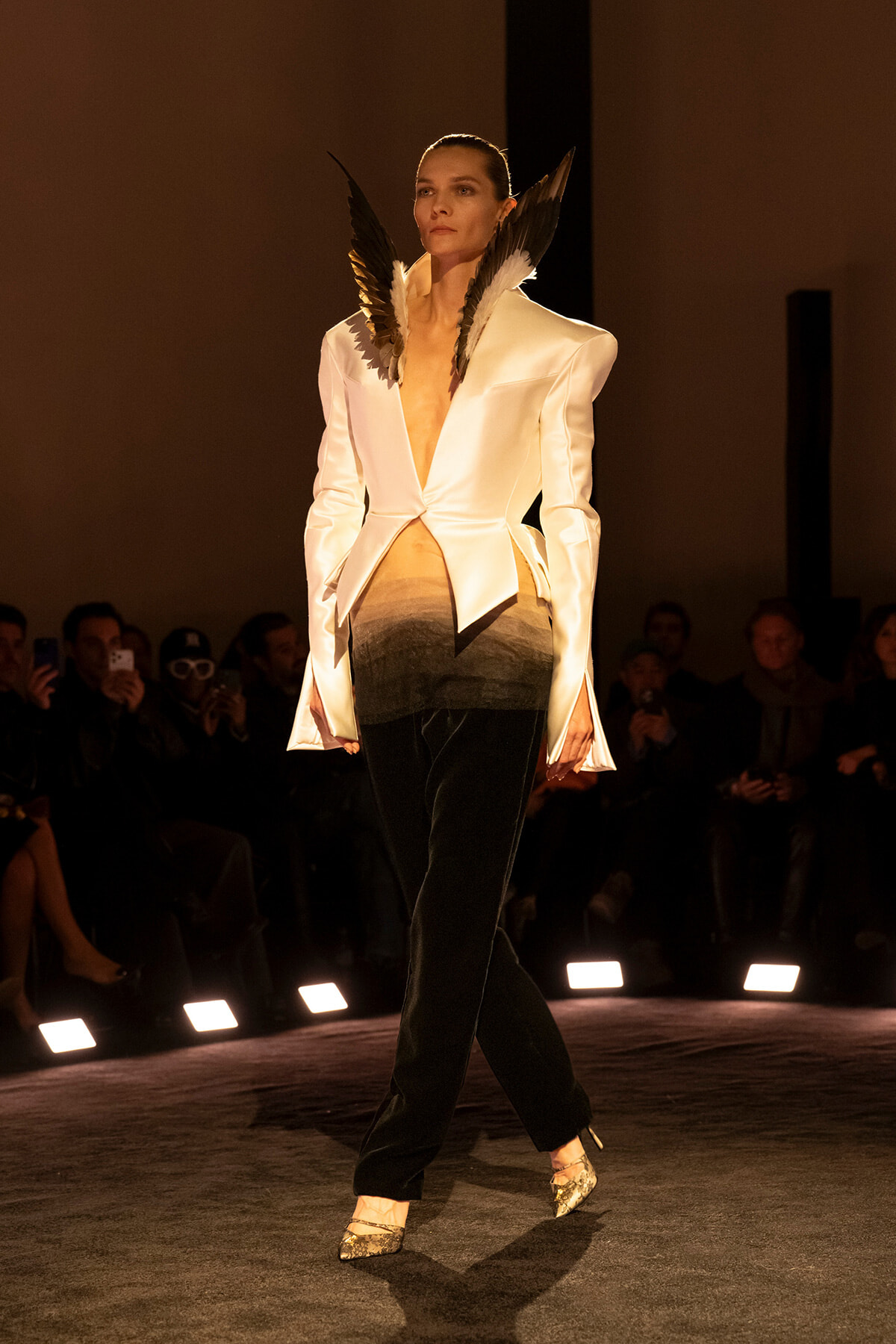 Model walks the runway in a cream tailored blazer with wing-like shoulder pieces, gradient pants, and metallic heels, audience in background.