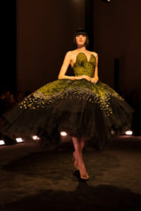 Model walks the runway in a dramatic emerald-green gown with a mossy-scale bodice and a voluminous black tulle skirt.