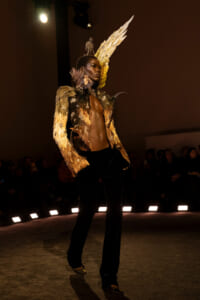 Model on a runway wearing a gold feathered jacket with large wing-like shoulders and a jeweled headpiece.
