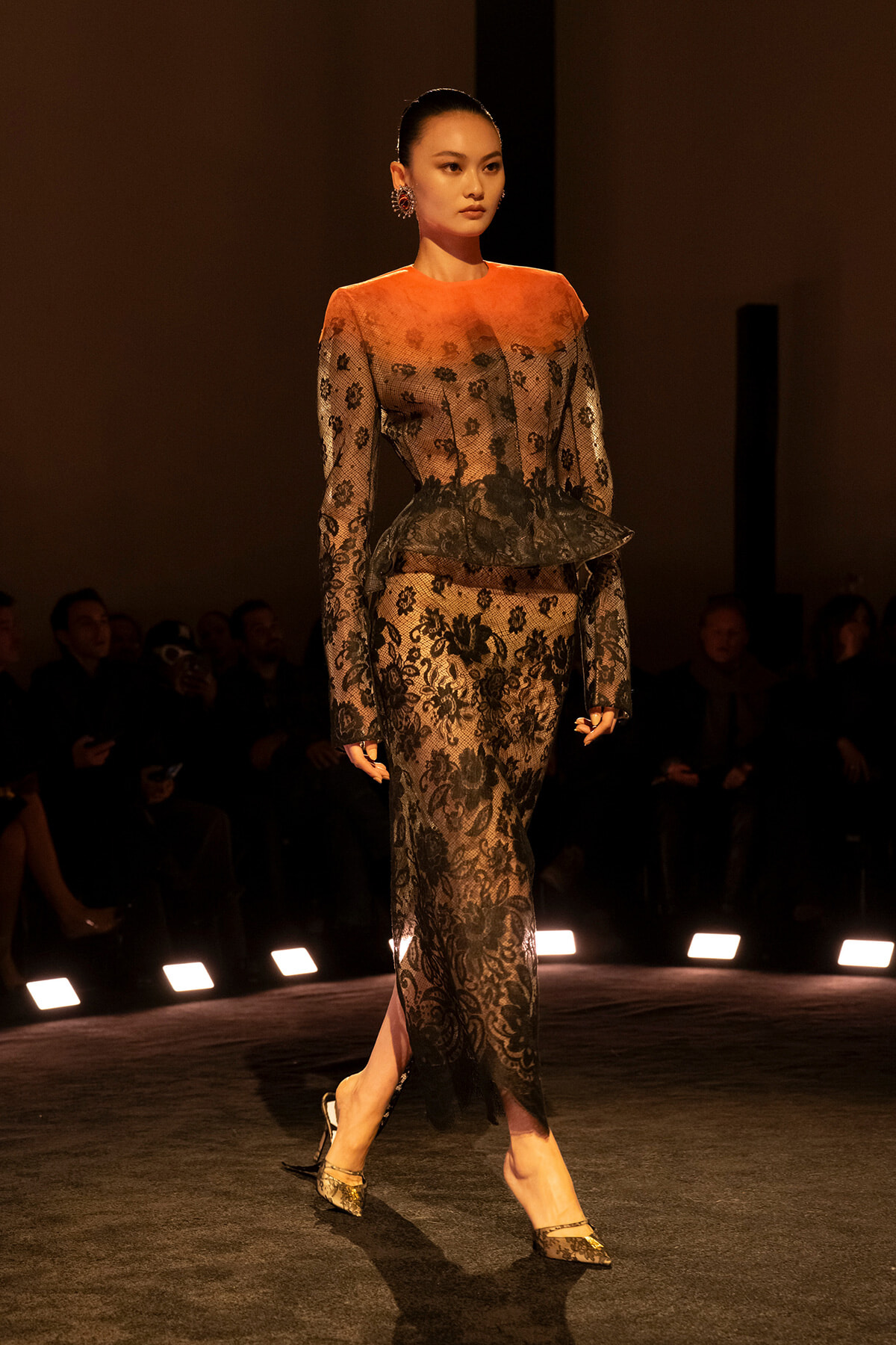 Model walks the runway in an orange-to-black lace gown with a peplum and statement earrings on a dim catwalk.