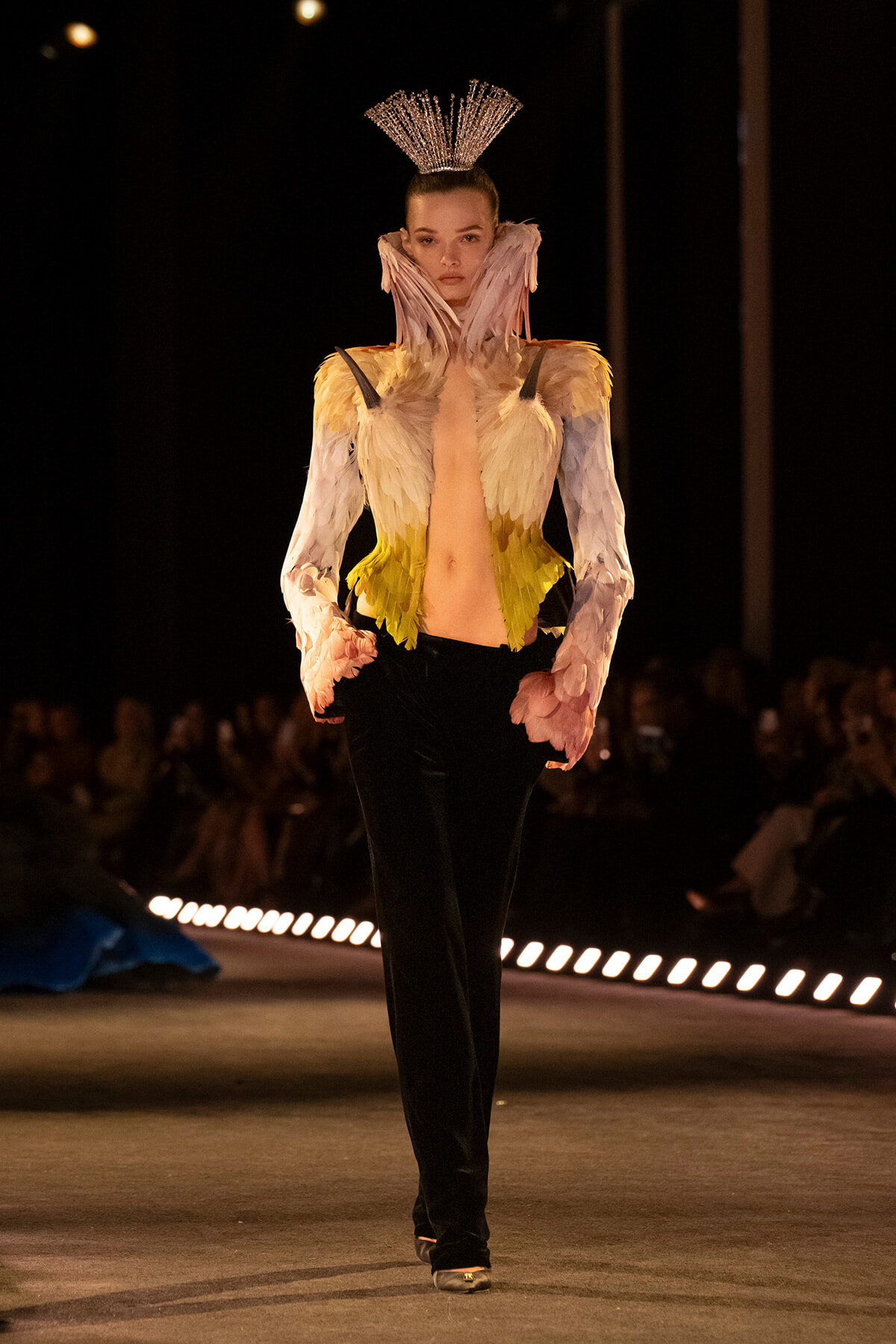 Model walks a runway in a dramatic feathered ensemble with a tall crown, pink–yellow feathered bodice and black pants