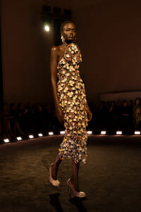 Model walks the runway in a one-shoulder couture dress covered in gold shell-like embellishments; audience watches from the sides under spotlighted stage lights.