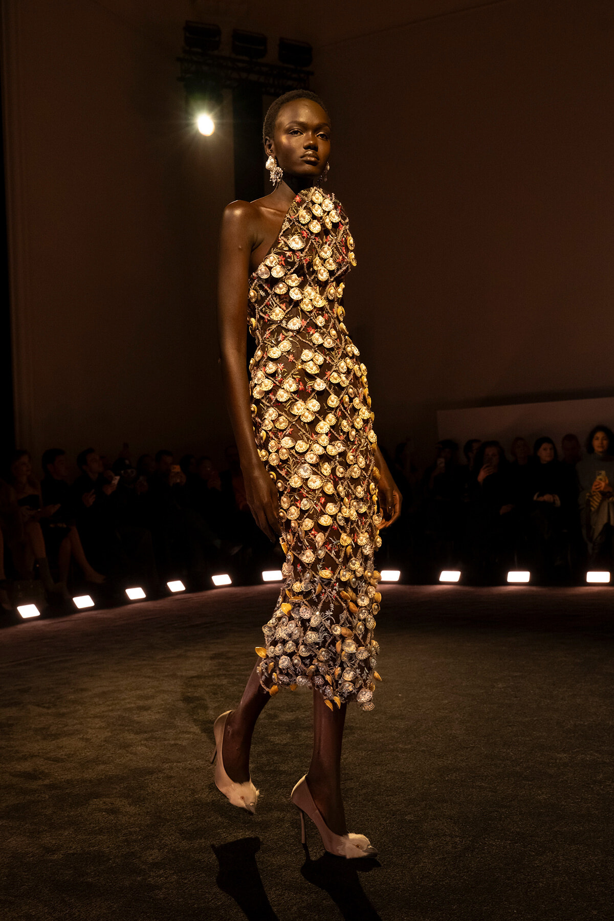 Model walks the runway in a one-shoulder couture dress covered in gold shell-like embellishments; audience watches from the sides under spotlighted stage lights.