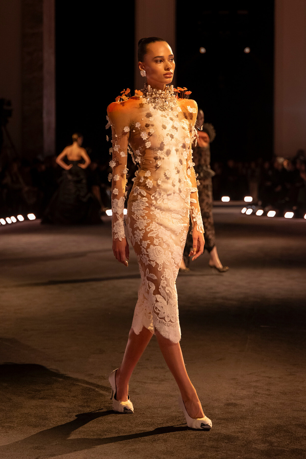 Runway model walks the catwalk in a sheer white lace midi dress adorned with floral appliqués and orange accents, paired with white heeled shoes.