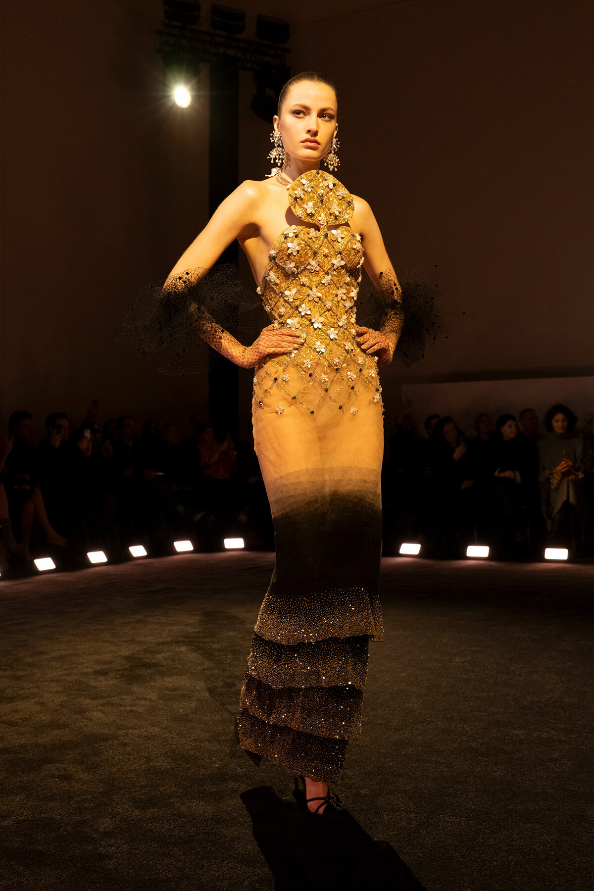 Model walks the runway in a gold, floral-embellished gown with sheer black lace sleeves and statement earrings under stadium lights.