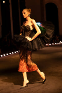 Model walks the runway in a strapless black tulle gown with a dramatic ruffled back and a lace skirt, wearing black heels.
