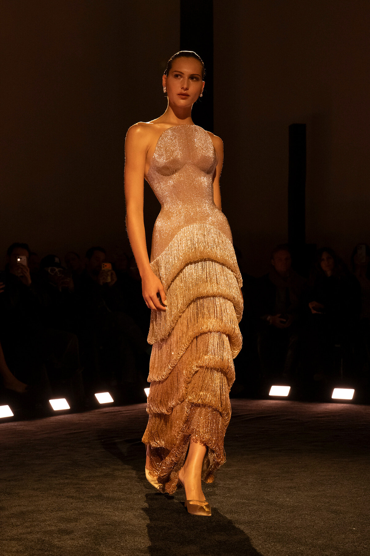 Model walking a runway in a shimmering champagne gown with tiered fringe layers and open shoulders, spotlighted against a dark background.
