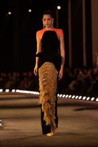 Model walks the runway in a black gown with orange shoulders and a gold beaded fringe skirt.