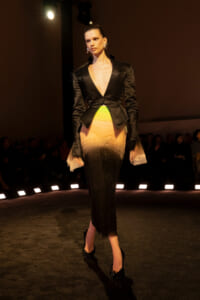 Model walking a fashion runway in a black, studded blazer with a plunging V neckline over a gradient dress fading from neon green to warm tan and black at the hem.