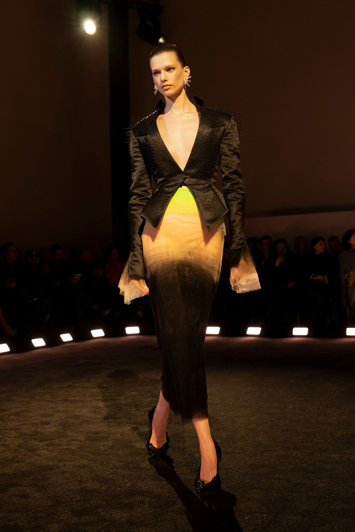 Model walking a fashion runway in a black, studded blazer with a plunging V neckline over a gradient dress fading from neon green to warm tan and black at the hem.