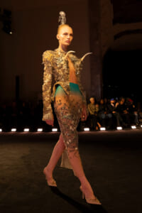Model walks the runway in a gold sequined jacket with horn accents and a sheer, teal-to-nude skirt, plus a crystal headpiece.
