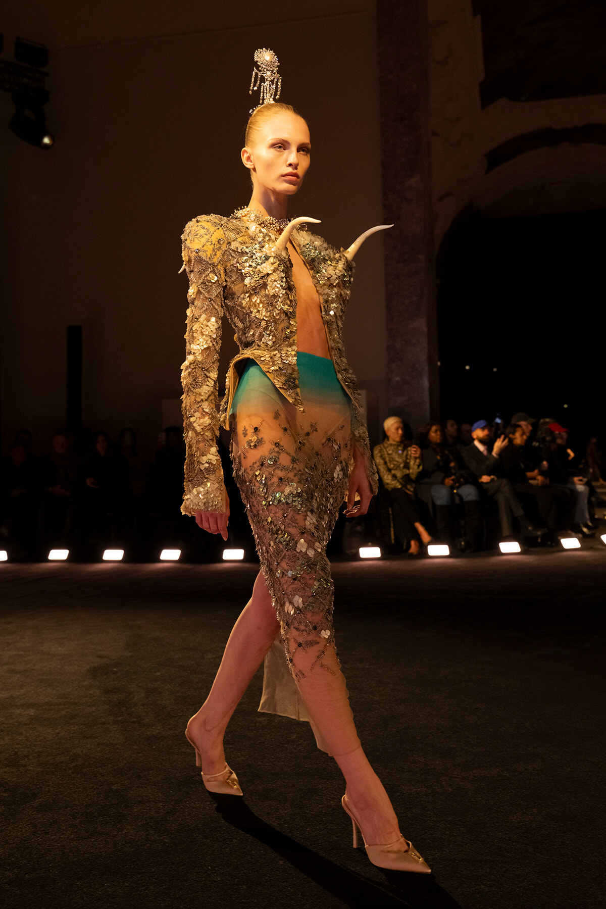 Model walks the runway in a gold sequined jacket with horn accents and a sheer, teal-to-nude skirt, plus a crystal headpiece.