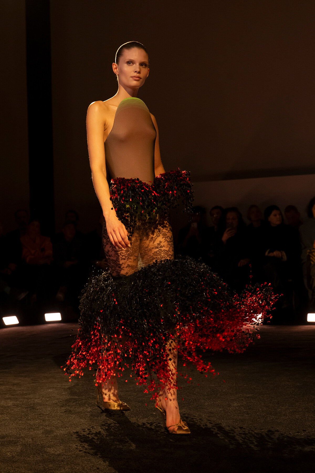 Model walks the runway in a sheer taupe bodysuit with a dramatic black-and-red feathered skirt.