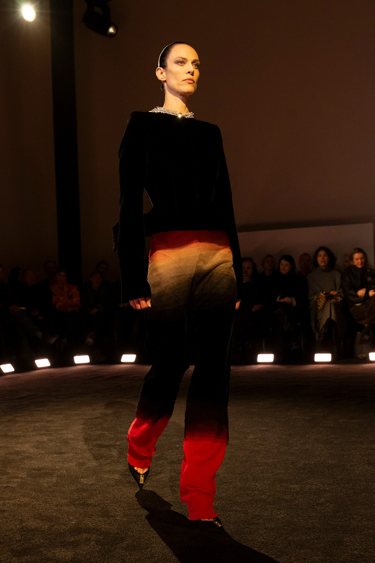 Full-length runway shot of a model in a black velvet top and gradient red-to-orange pants, audience in dim seating under warm lights.