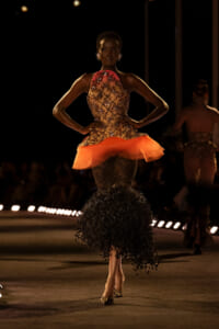 Model on a dim runway wearing a beaded, embroidered bodice gown with an orange sheer peplum and a black fringe mermaid skirt under spotlights on a dark catwalk.