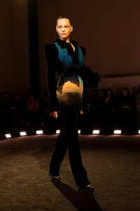 Model walking a runway in a black outfit with teal feather accents and a gradient skirt, spotlight on stage, audience in the background.