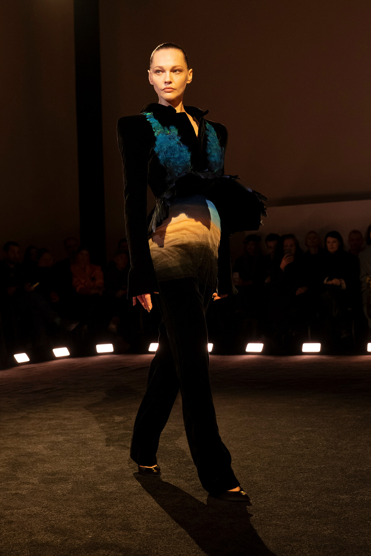Model walking a runway in a black outfit with teal feather accents and a gradient skirt, spotlight on stage, audience in the background.