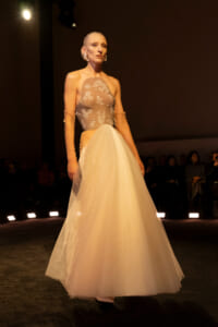 Model walking a bridal-inspired gown down the runway, featuring a sheer lace bodice and full pale tulle skirt, under warm spotlight.