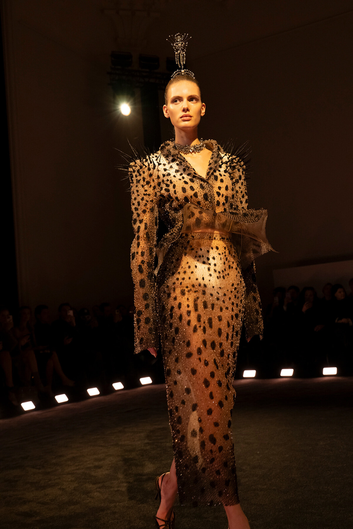 Model walks a runway in a sheer, leopard-spotted gown with spiky shoulders and a tall beaded headpiece, under warm spotlight.