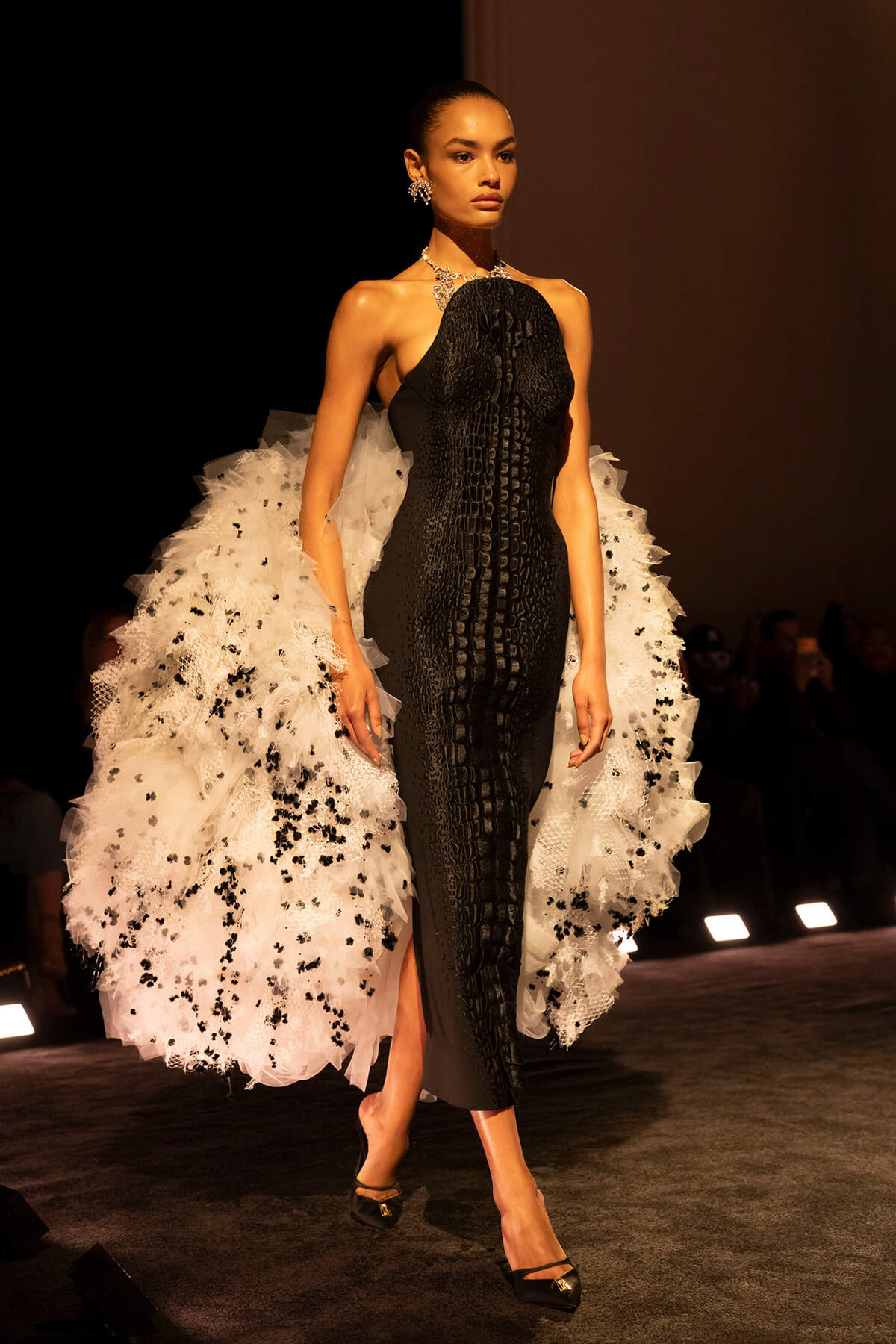 Model walking a runway in a black textured halter dress with a dramatic white tulle train speckled in black, wearing statement jewelry.