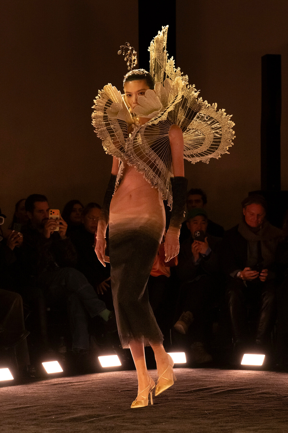 Model on a runway wearing an oversized, intricate lace sculptural collar with a gradient dress; audience seated in dim lighting.