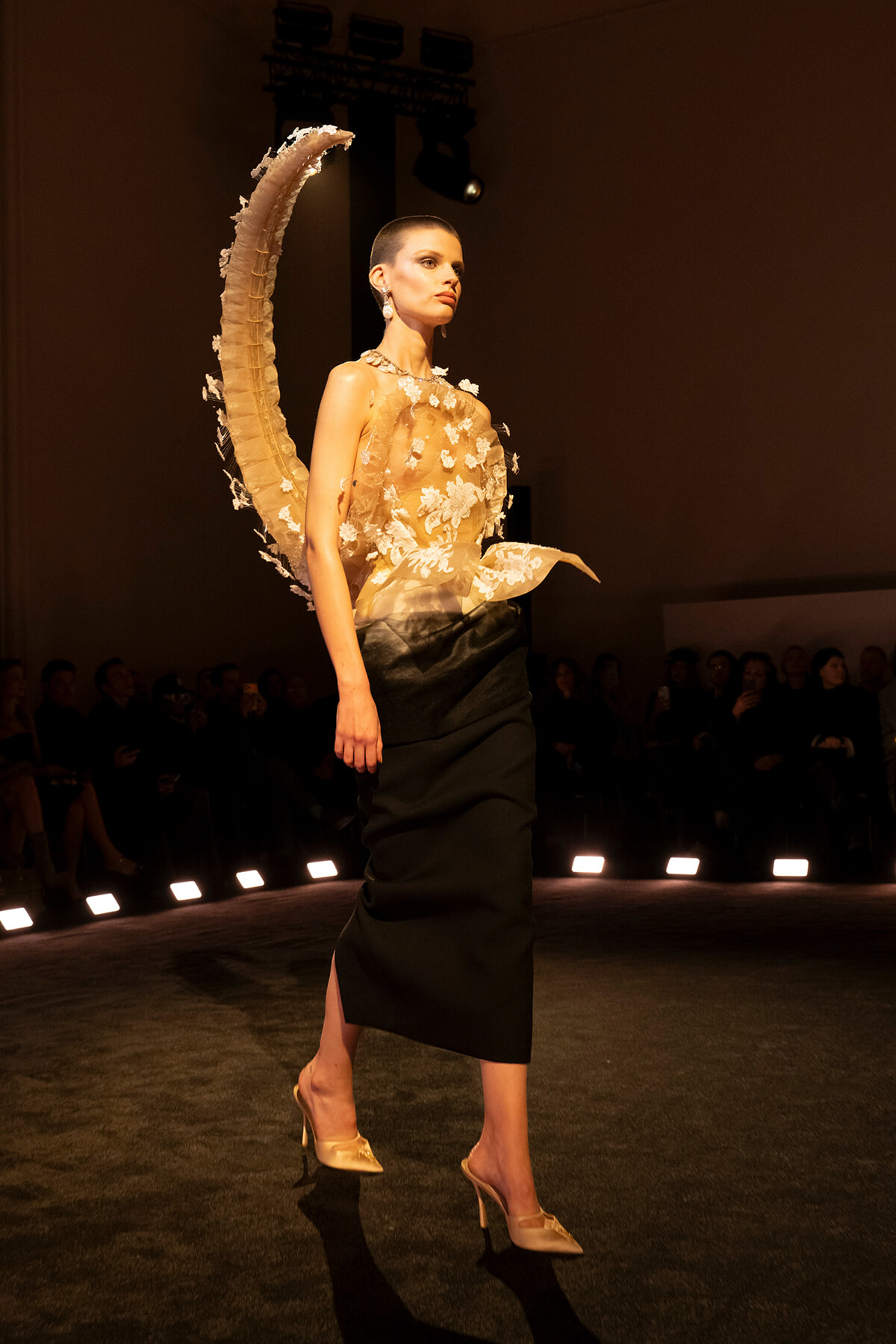 Model walks a runway in a sheer beige floral top and black midi skirt, with a large curved wing-like accessory behind her.