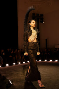 Model walks the runway in a black lace ensemble with a high-neck lace top and long skirt, dramatic shoulder embellishment, and vintage gloves.