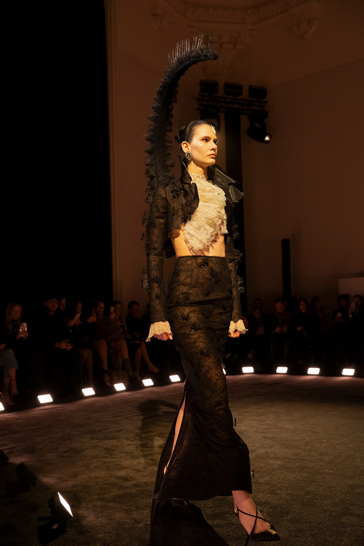 Model walks the runway in a black lace ensemble with a high-neck lace top and long skirt, dramatic shoulder embellishment, and vintage gloves.
