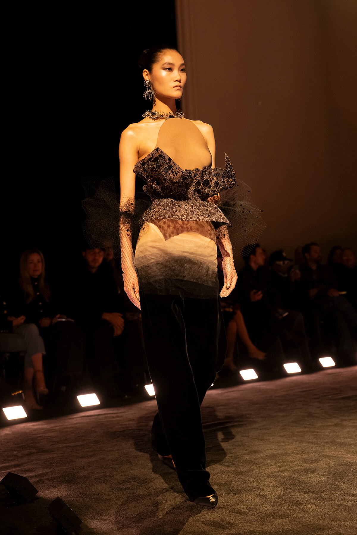 Model walks a fashion runway in a dramatic black and nude gown with ruffled lace bodice, net sleeves, and statement jewelry, under warm stage lighting.