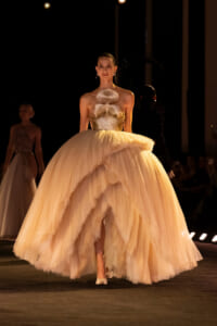 Model on a runway wearing a voluminous peach tulle ball gown with a fluffy feathered bodice.