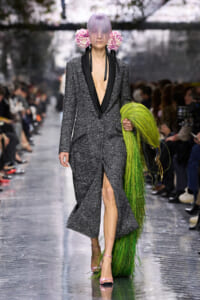 Runway model in a textured gray coat with black lapels and deep V, pink heels, and a bright green fringe prop; lavender hair with pink floral headpiece